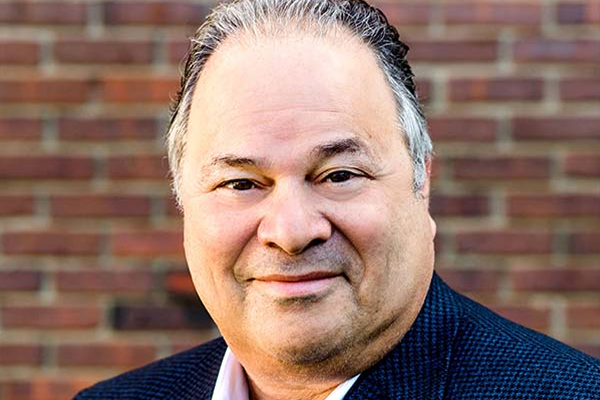 Smiling man with a light shirt and dark blazer, standing against a brick wall background.