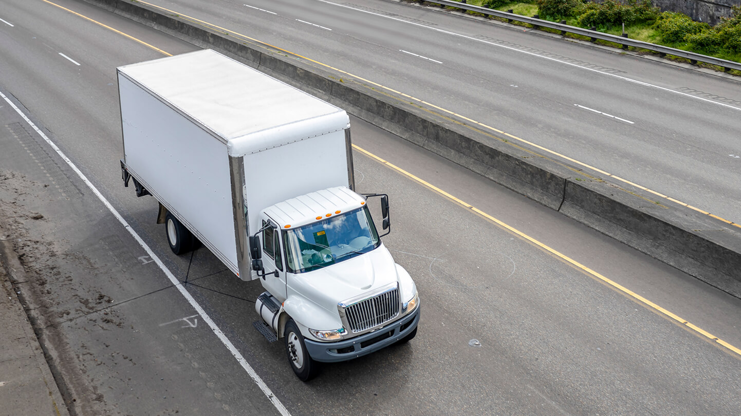 White box truck driving on an empty, multi-lane highway during the day, surrounded by greenery.