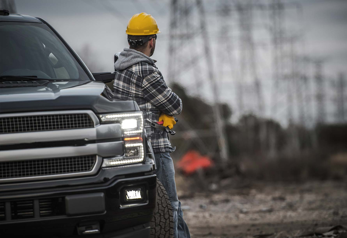 A construction worker in a helmet leans on a truck, looking at an overcast sky and power lines in a barren area.
