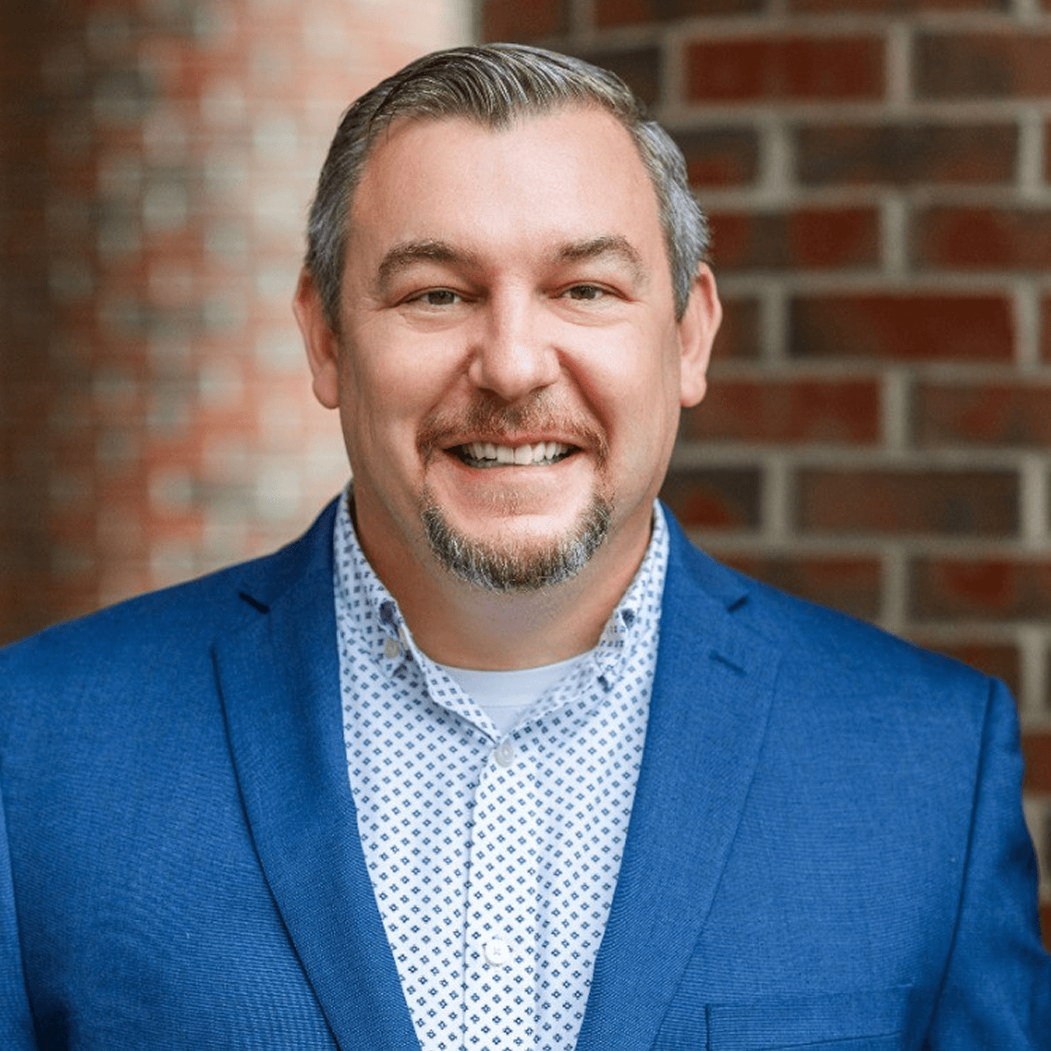 Smiling man in a blue blazer and patterned shirt in front of a brick wall.